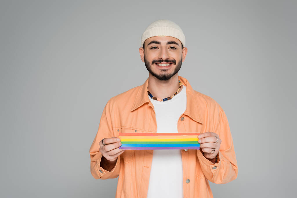 Veselý homosexuál držící vlajku lgbt a dívající se na kameru izolovanou na šedi   - Fotografie, Obrázek