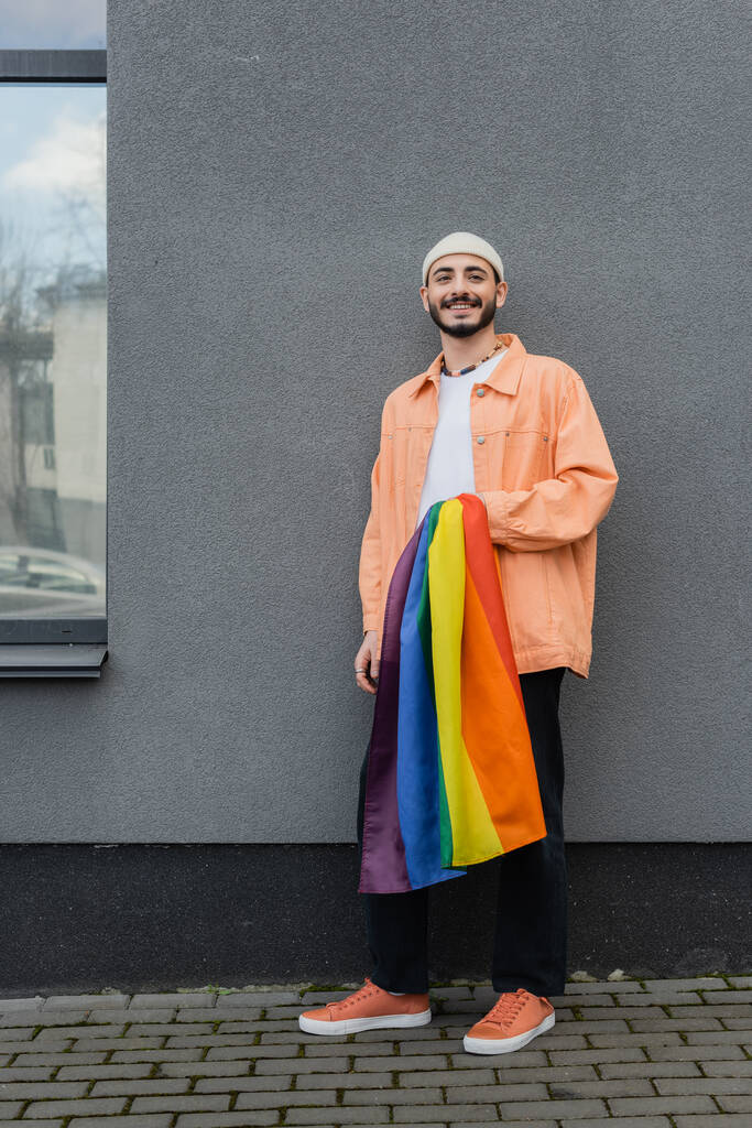 sonriente gay hombre holding lgbt bandera cerca de edificio al aire libre  - Foto, Imagen