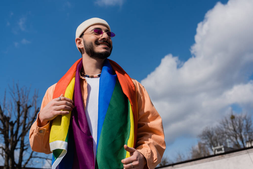 Низкий угол обзора улыбающегося гомосексуалиста в солнцезащитных очках с флагом lgbt на городской улице, Международный день борьбы с гомофобией - Фото, изображение