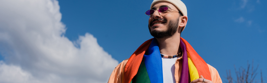Низкий угол зрения беззаботного гея в солнцезащитных очках с флагом lgbt, стоящим на улице, баннер  - Фото, изображение