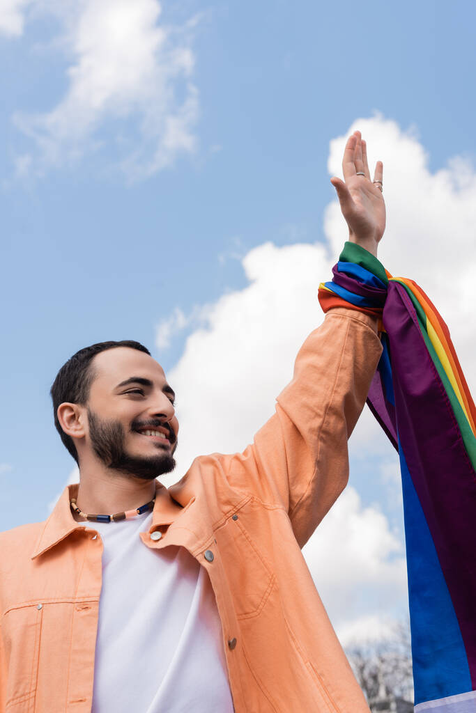 Fröhlicher schwuler Mann mit lgbt-Fahne schwenkt Hand auf urbaner Straße, Internationaler Tag gegen Homophobie - Foto, Bild