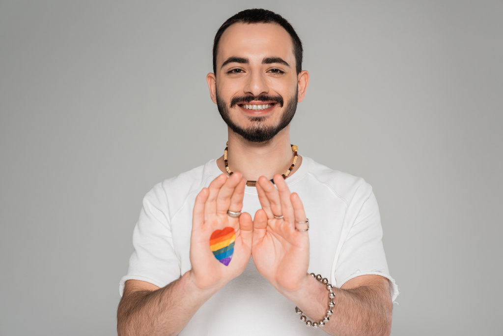 Young smiling gay man with lgbt flag on hand looking at camera isolated on grey, International day against homophobia - Photo, Image