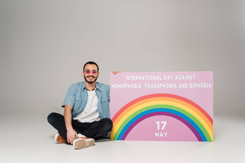 allegro gay uomo vicino cartello con internazionale giornata contro omofobia, transfobia e bifobia lettering su grigio  - Foto, immagini