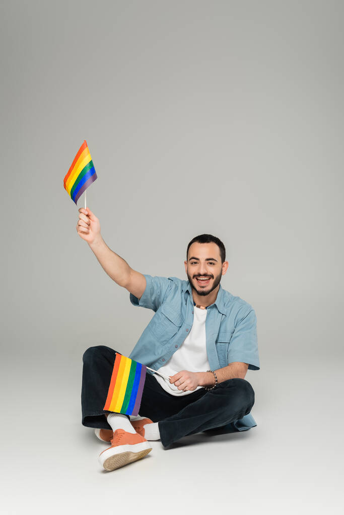 Улыбающийся гей с флагами, сидящий на сером фоне  - Фото, изображение