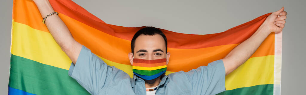 Bruneta gayové v lékařské masce drží vlajku lgbt izolované na šedé, prapor  - Fotografie, Obrázek