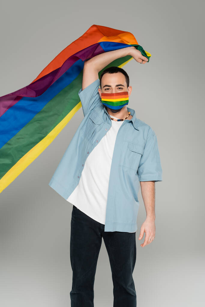 Nuori homoseksuaali mies lääketieteellisessä naamiossa pitämässä lgbt-lippua eristettynä harmaana, kansainvälisenä homofobiapäivänä   - Valokuva, kuva