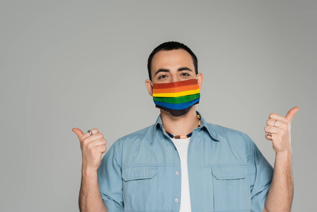 Morena homossexual homem em máscara médica em cores lgbt mostrando como gesto isolado no cinza, Dia Internacional Contra a Homofobia - Foto, Imagem