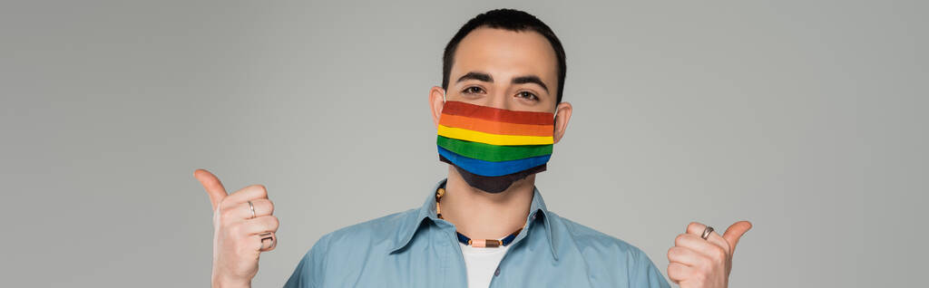 Νεαρός ομοφυλόφιλος άνδρας με ιατρική μάσκα με σημαίες Igbt που δείχνουν τους αντίχειρες επάνω απομονωμένους στο γκρι, banner  - Φωτογραφία, εικόνα