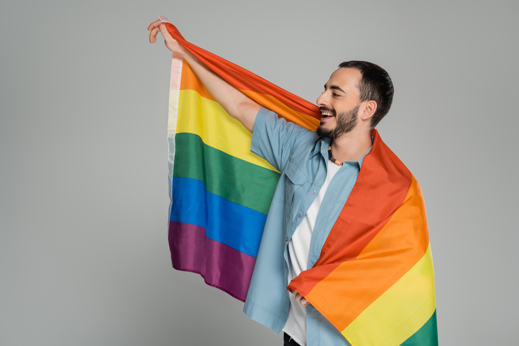Hombre gay barbudo y alegre sosteniendo bandera lgbt y mirando hacia otro lado aislado en gris, Día Internacional contra la Homofobia - Foto, imagen