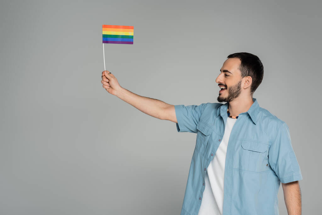 Вид збоку веселий гомосексуальний чоловік тримає прапор lgbt і дивиться ізольовано на сірий
   - Фото, зображення