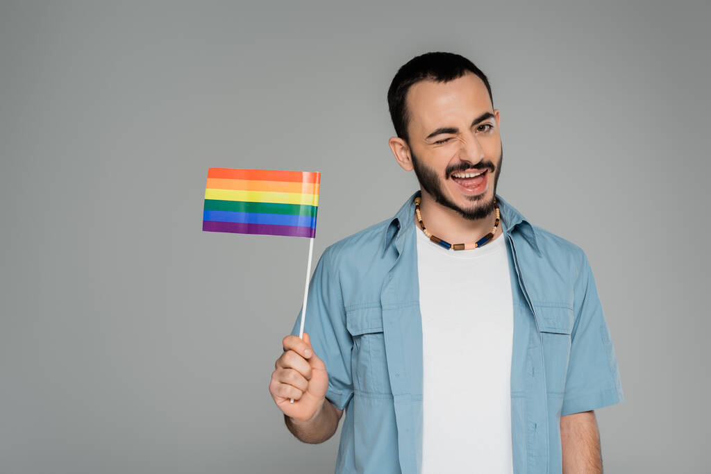 Uomo omosessuale spensierato che tiene la bandiera lgbt e strizza l'occhio alla telecamera isolata sul grigio, Giornata internazionale contro l'omofobia - Foto, immagini