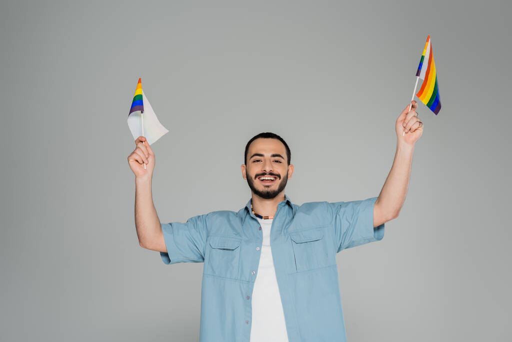Young and cheerful gay man holding lgbt flags and looking at camera isolated on grey   - Photo, Image