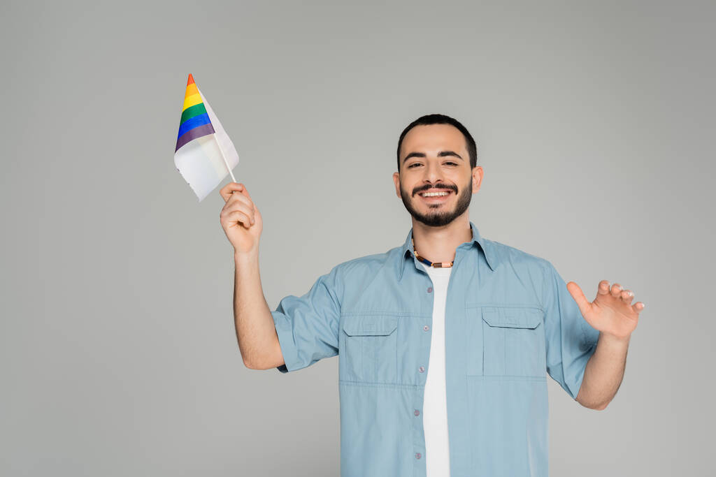 Bärtiger schwuler Mann im Hemd lächelt und hält Regenbogenfahne isoliert auf grau   - Foto, Bild