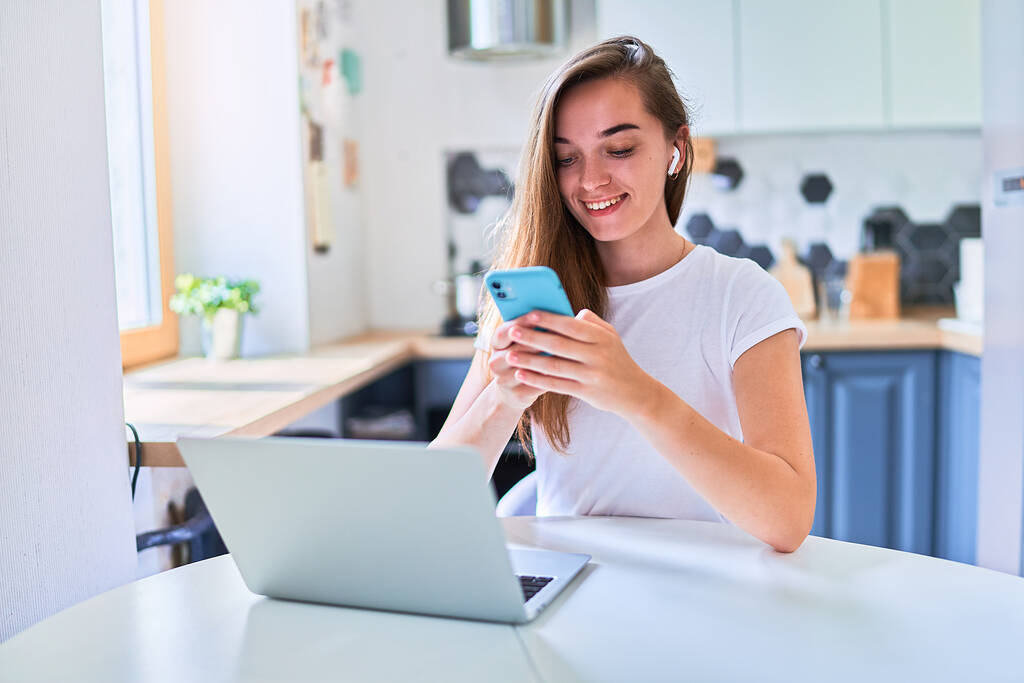 Młoda atrakcyjna uśmiechnięta szczęśliwa millenium dziewczyna siedzi przy stole i za pomocą laptopa i telefonu do czatowania online, surfingu internetowego, przeglądania i sieci społecznych w domu  - Zdjęcie, obraz