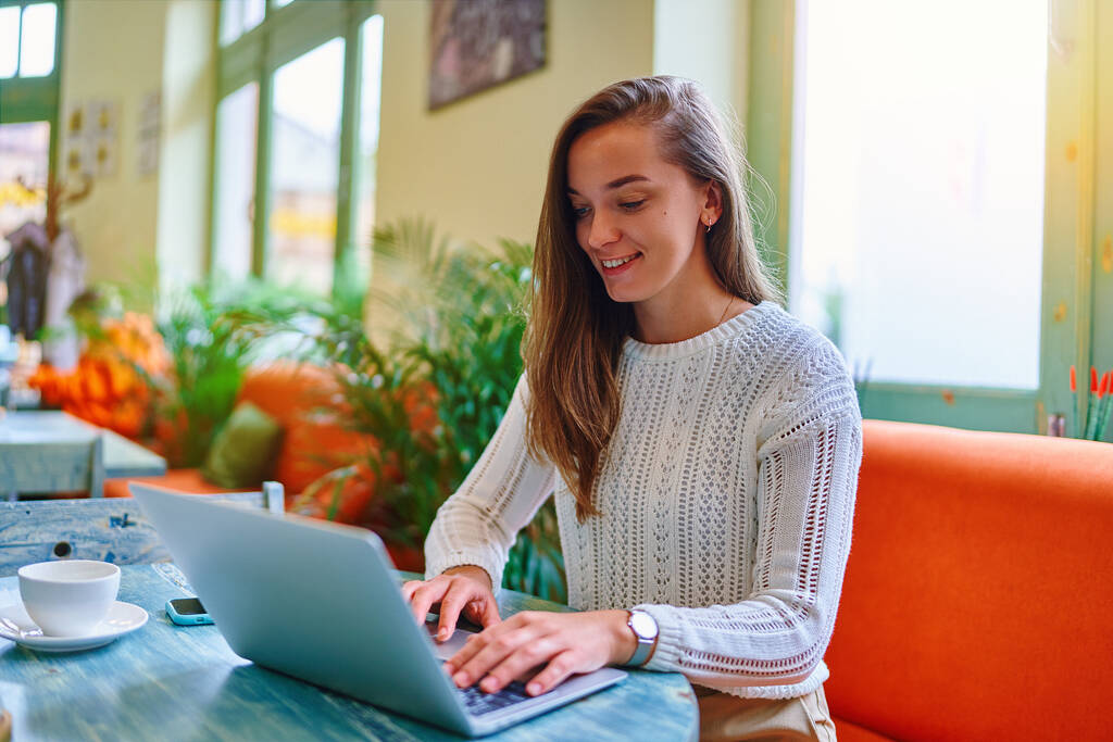Портрет милой молодой счастливой улыбающейся умной современной копирайтер внештатной девушки, печатающей на клавиатуре ноутбука во время работы в Интернете удаленно в кафе  - Фото, изображение
