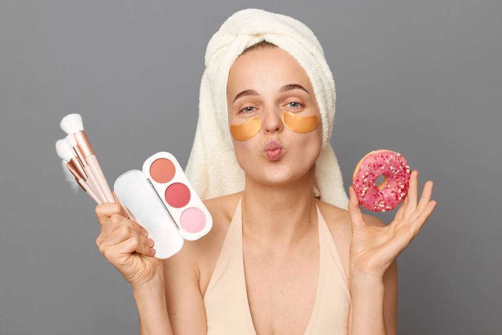 Mooie grappige jonge vrouw met vlekken onder de ogen gewikkeld in handdoek poseren geïsoleerd over grijze achtergrond het verzenden van lucht kussen terwijl het doen van make-up en het eten van donut. - Foto, afbeelding