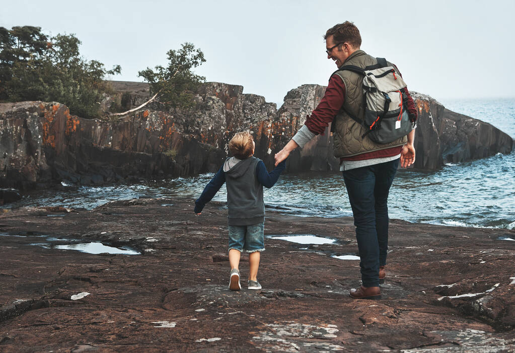 Skákat celou cestu domů. Zadní pohled záběr veselého otce a syna držící se za ruce při chůzi u oceánu venku během dne - Fotografie, Obrázek