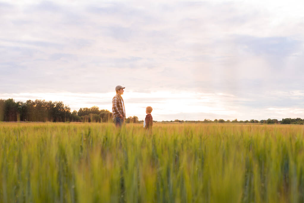Farmer and his son in front of a sunset agricultural landscape. Man and a boy in a countryside field. The concept of fatherhood, country life, farming and country lifestyle. - 写真・画像