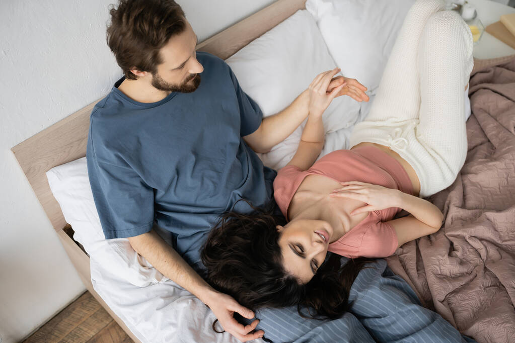 Top näkymä parrakas mies koskettaa käsi hymyilevä brunette tyttöystävä sängyssä kotona  - Valokuva, kuva
