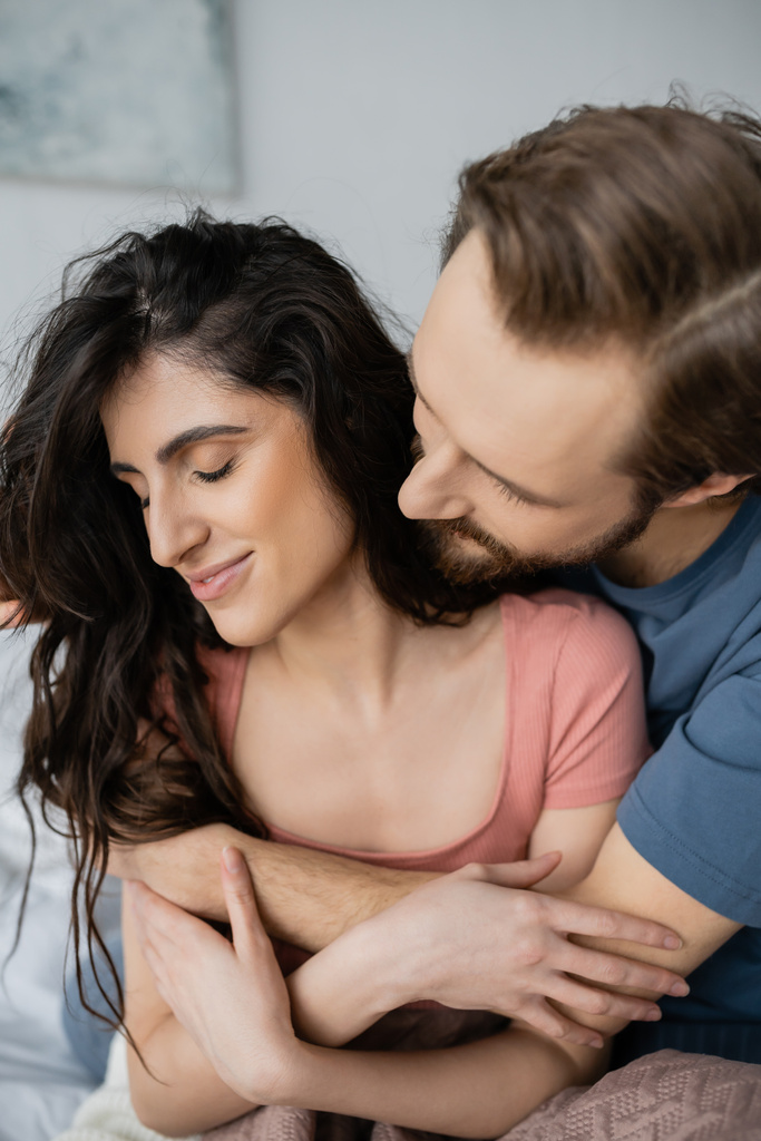 Bearded man hugging smiling brunette girlfriend in bedroom  - Photo, Image