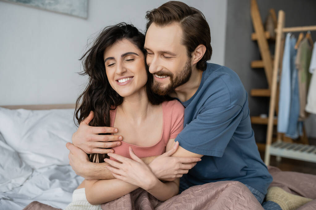 Веселый мужчина обнимает брюнетку подружку в пижаме на кровати дома  - Фото, изображение