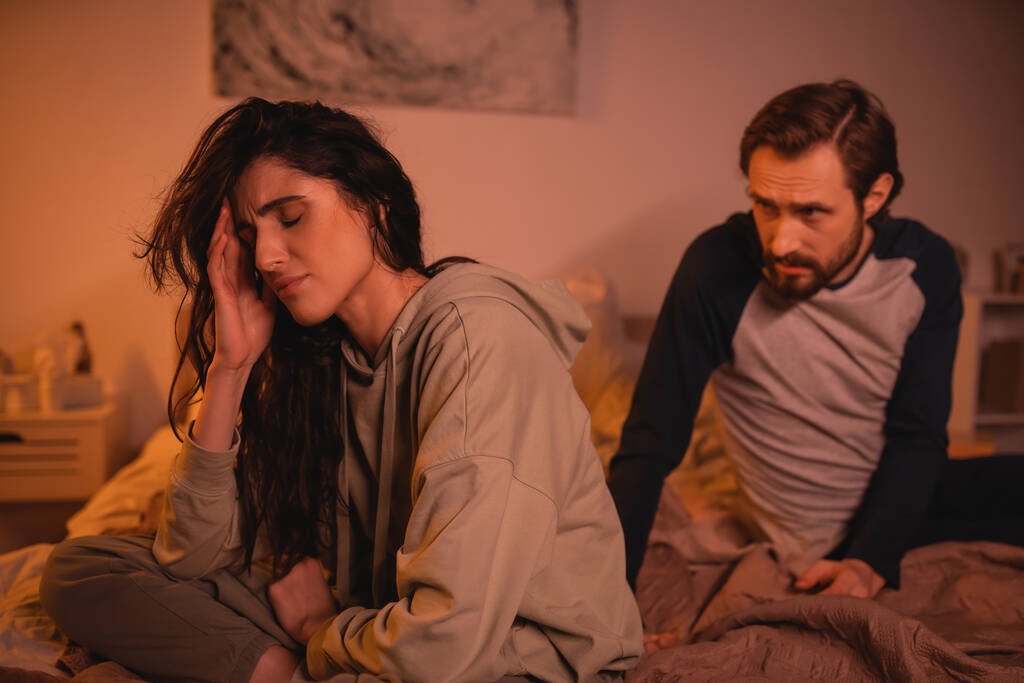 Расстроенная женщина, сидящая рядом с расплывчатым бойфрендом и разговаривающая по вечерам в постели  - Фото, изображение