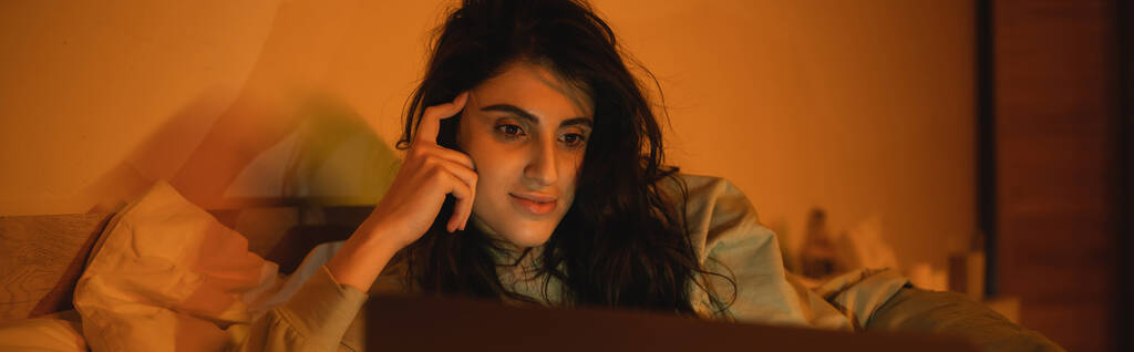 Μελαχρινή γυναίκα βλέποντας ταινία σε θολή φορητό υπολογιστή στο υπνοδωμάτιο τη νύχτα, πανό  - Φωτογραφία, εικόνα