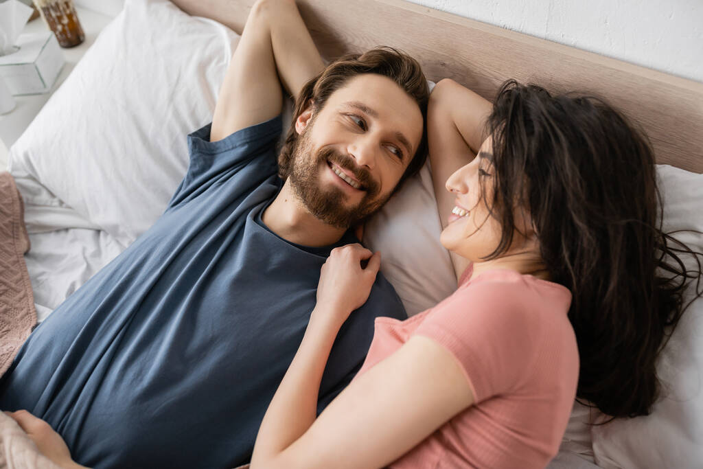 Вид зверху усміхненої брюнетки жінка дивиться на бородатого хлопця на ліжко вранці
  - Фото, зображення