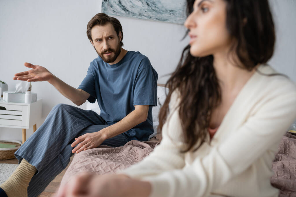 Hombre ofendido en pijama hablando con novia borrosa mientras está sentado en la cama en casa  - Foto, Imagen