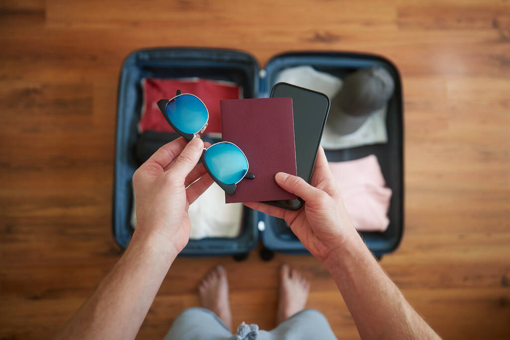 Imballaggio valigia e pianificazione viaggio avventura estiva. Uomo con in mano smart phone, passaporto e occhiali da sole sopra il bagaglio aperto - Foto, immagini