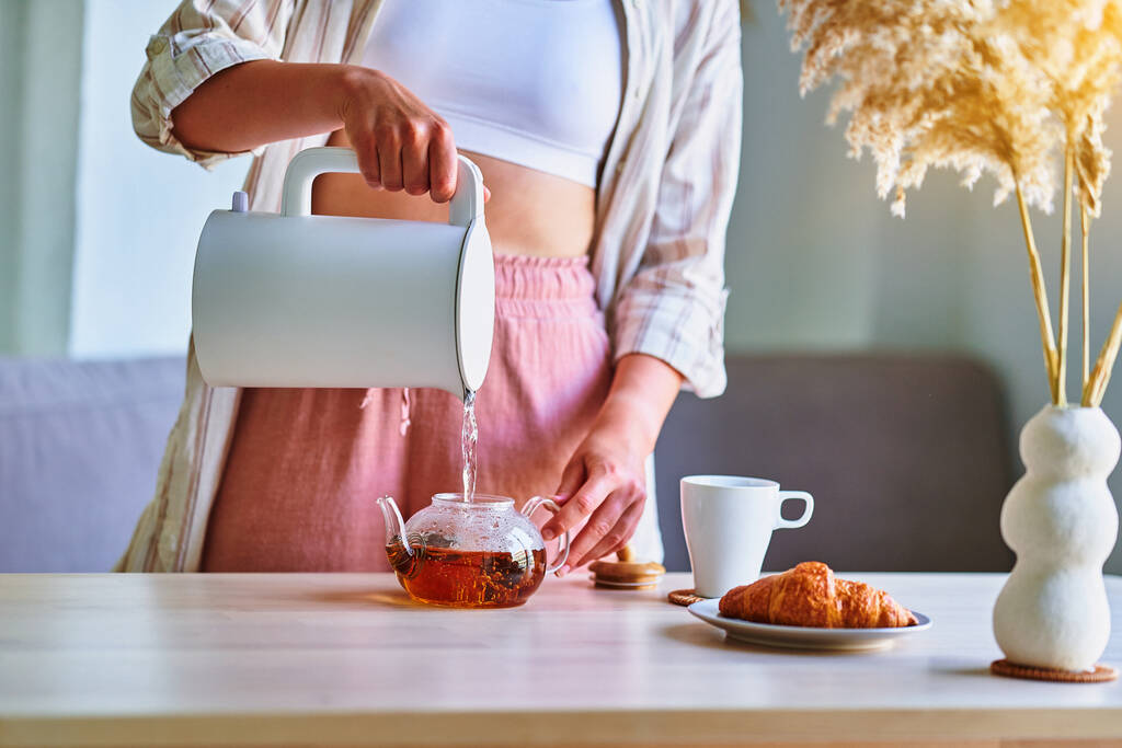 Kadın evde çay demlemek için modern elektrikli çaydanlıktan bir fincana kaynar su döküyor.  - Fotoğraf, Görsel