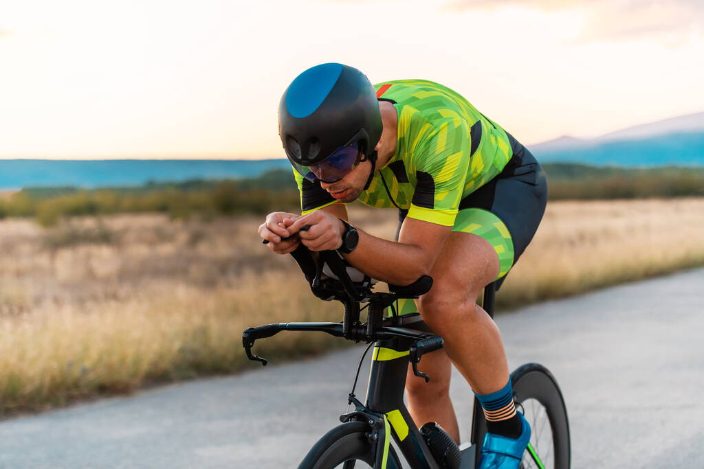  Zamknij zdjęcie triatlonisty jadącego na rowerze podczas zachodu słońca, przygotowującego się do maratonu. Ciepłe kolory nieba zapewniają piękne tło dla jego zdecydowanego i skoncentrowanego wysiłku - Zdjęcie, obraz