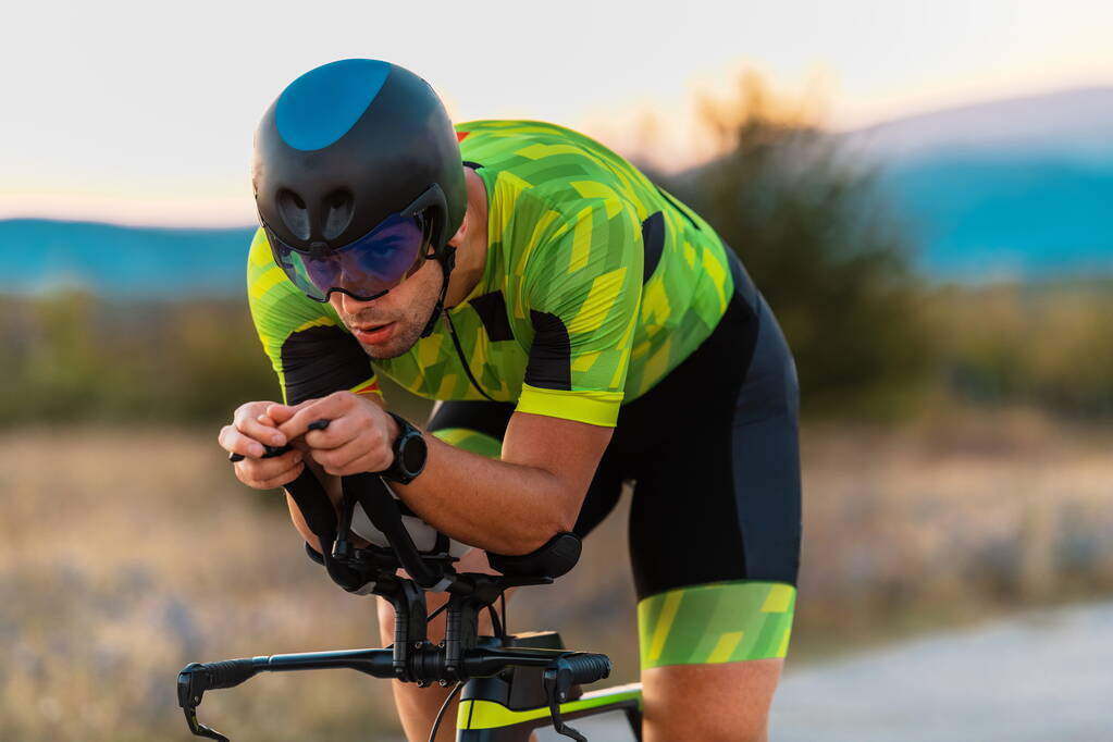  Photo rapprochée d'un triathlète en vélo au coucher du soleil, se préparant pour un marathon. Les couleurs chaudes du ciel fournissent une belle toile de fond pour son effort déterminé et concentré - Photo, image