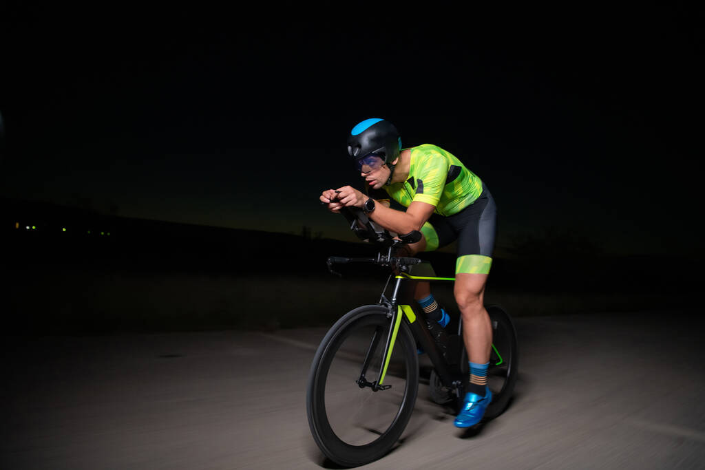 Ein Triathlet fährt in der Dunkelheit der Nacht mit dem Fahrrad, um sich auf einen Marathon vorzubereiten. Der Kontrast zwischen Dunkelheit und Licht seines Fahrrads erzeugt ein Gefühl von Dramatik und unterstreicht die - Foto, Bild