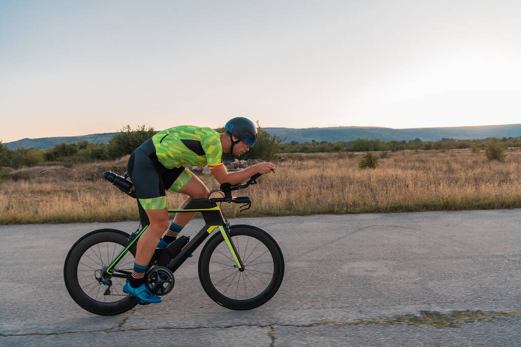  Triathlet auf seinem Fahrrad bei Sonnenuntergang, um sich auf einen Marathon vorzubereiten. Die warmen Farben des Himmels bieten eine schöne Kulisse für seinen entschlossenen und konzentrierten Einsatz - Foto, Bild
