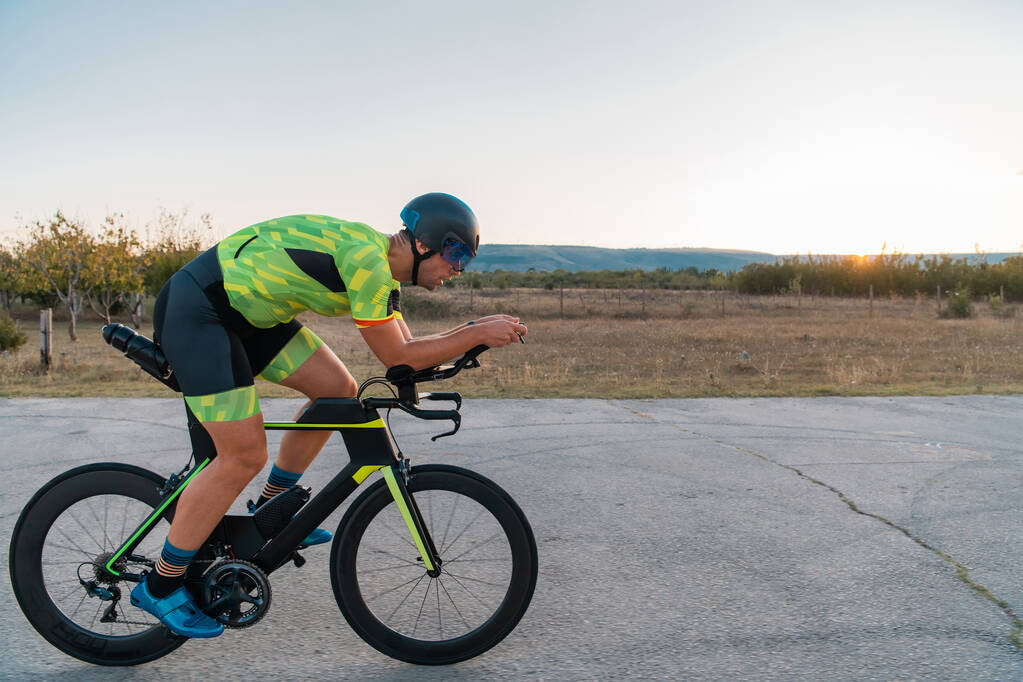  Triatleet fietst tijdens zonsondergang, bereidt zich voor op een marathon. De warme kleuren van de lucht bieden een prachtige achtergrond voor zijn vastberaden en gerichte inspanning - Foto, afbeelding
