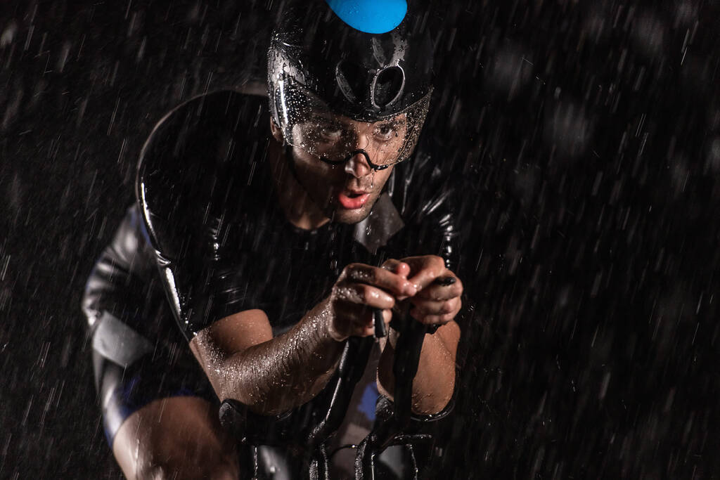 Een triatleet die de regen trotseert terwijl hij door de nacht fietst, en zich voorbereidt op de komende marathon. De wazige regendruppels op de voorgrond en de donkere, humeurige sfeer op de achtergrond voegen toe - Foto, afbeelding