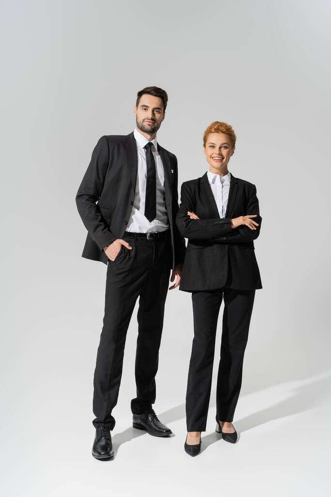 longitud completa de colegas de negocios sonrientes en negro y elegante ropa formal sobre fondo gris - Foto, imagen