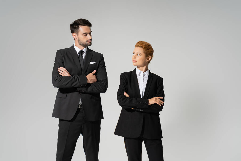 skeptical business people in black pantsuits standing with folded arms and looking at each other isolated on grey - Photo, Image