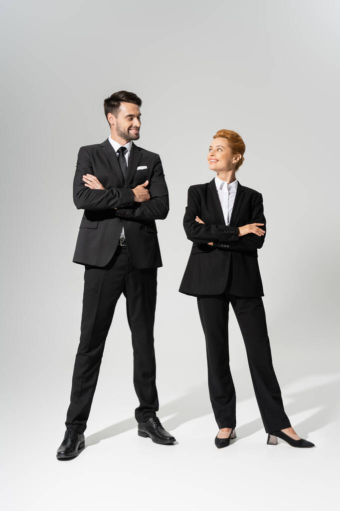 in voller Länge auf gut gelaunten Geschäftskollegen in stylischen Strumpfhosen, die einander anlächeln, während sie mit verschränkten Armen vor grauem Hintergrund stehen - Foto, Bild