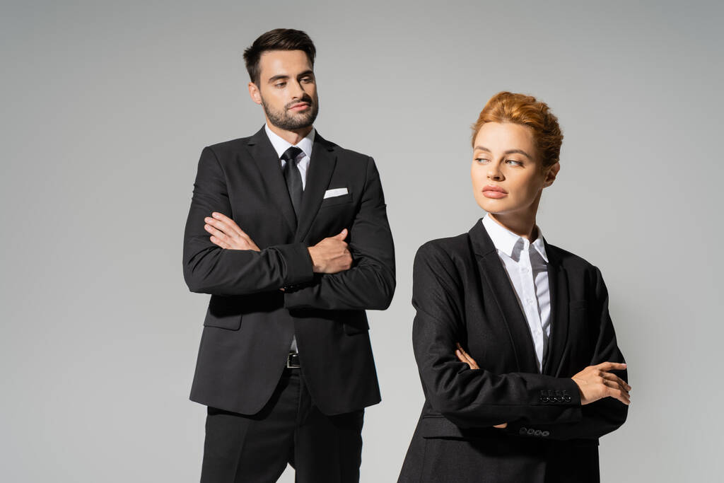 skeptischer Geschäftsmann im schicken Anzug, der die rothaarige Frau in formeller Kleidung isoliert auf grau betrachtet - Foto, Bild