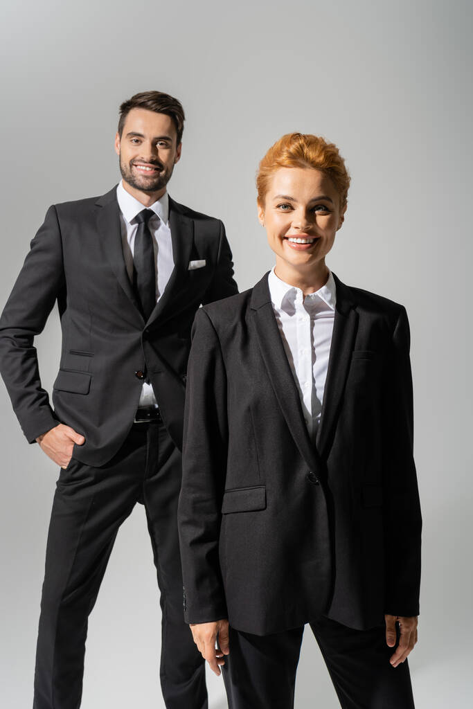 cheerful redhead businesswoman looking at camera near smiling colleague standing with hand in pocket isolated on grey - Photo, Image