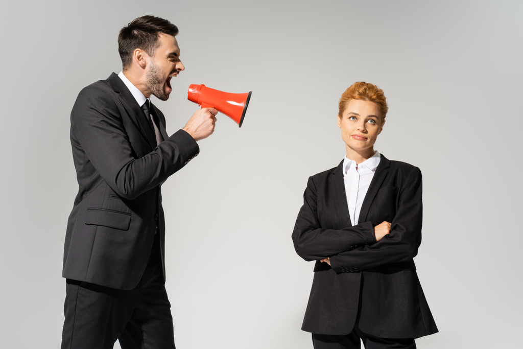 angry businessman screaming in loudspeaker near skeptical woman standing with crossed arms isolated on grey - Photo, Image