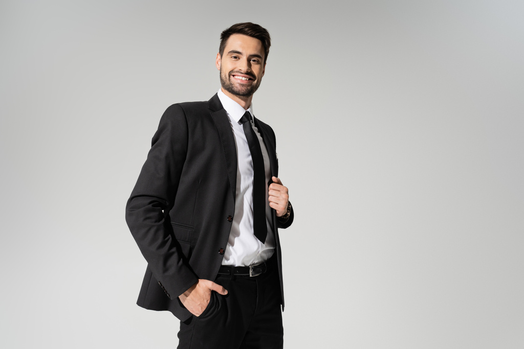 γενειοφόρος επιχειρηματίας με μαύρο κοστούμι στέκεται με το χέρι στην τσέπη και χαμογελώντας στην κάμερα απομονώνονται σε γκρι - Φωτογραφία, εικόνα