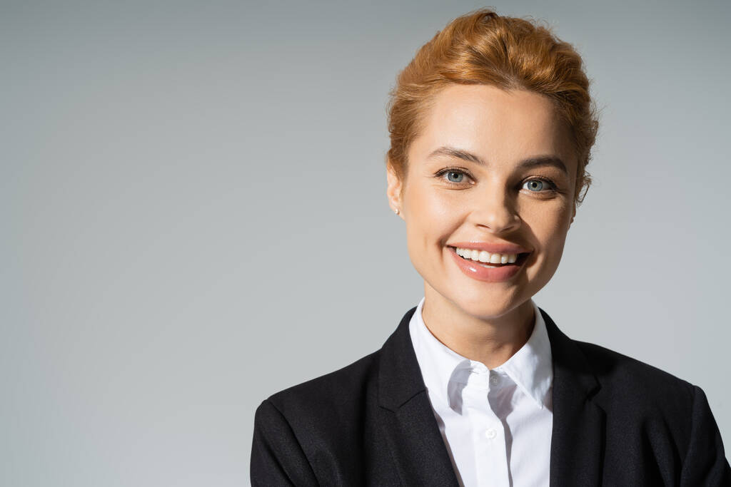 portret rozweselonej bizneswoman z rudymi włosami uśmiechniętej do kamery odizolowanej na szarości - Zdjęcie, obraz