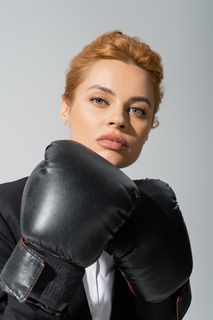 retrato de mujer de negocios pelirroja confiada en guantes de boxeo mirando a la cámara aislada en gris - Foto, imagen