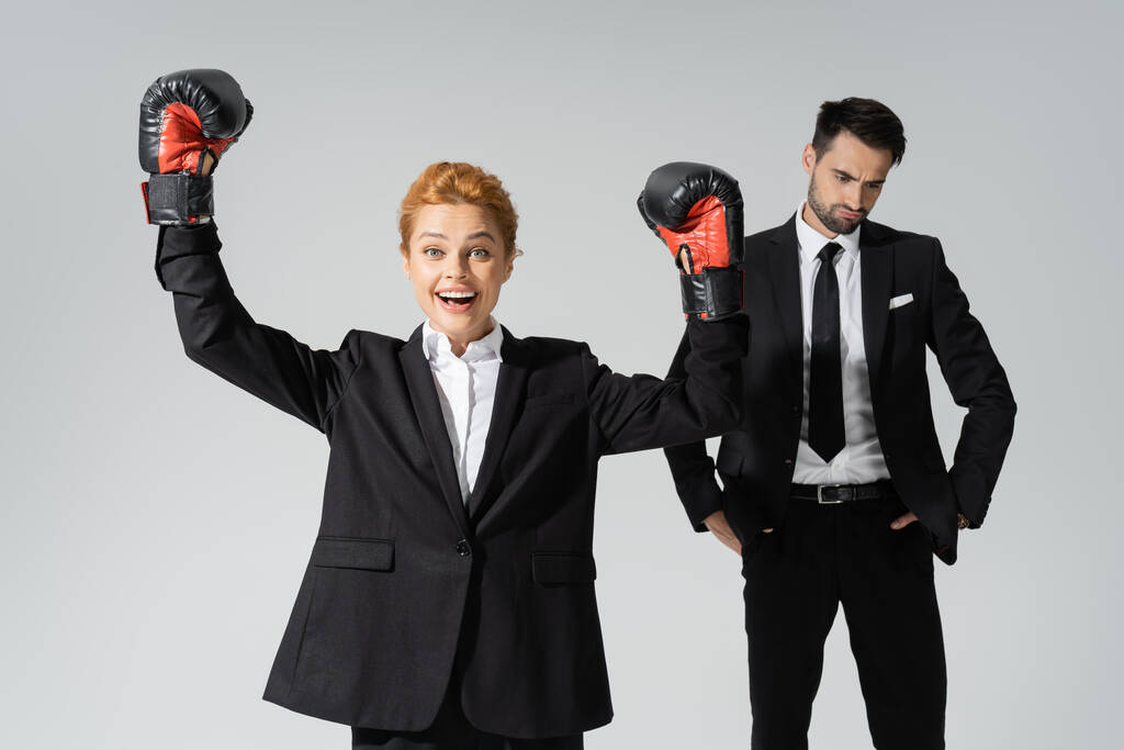 περιχαρής επιχειρηματίας σε γάντια πυγμαχίας δείχνει κερδίσει χειρονομία κοντά αναστατωμένος επιχειρηματικό ανταγωνιστή απομονώνονται σε γκρι - Φωτογραφία, εικόνα