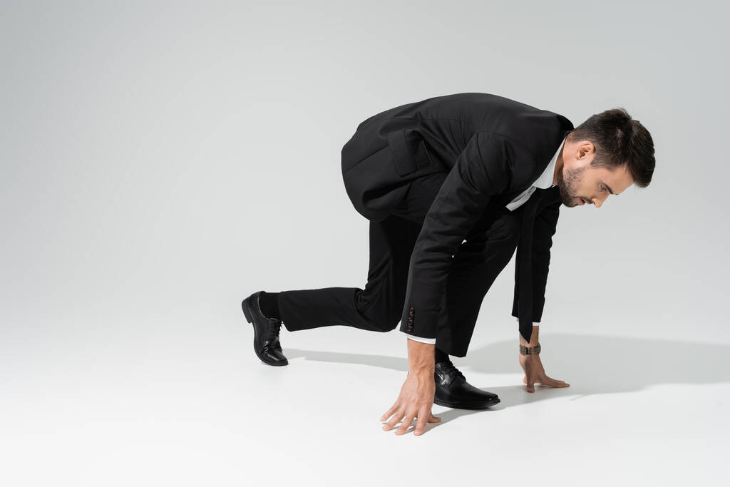 πλαϊνή άποψη του επιχειρηματία με μαύρο κοστούμι στέκεται σε χαμηλή θέση εκκίνησης σε γκρι φόντο - Φωτογραφία, εικόνα