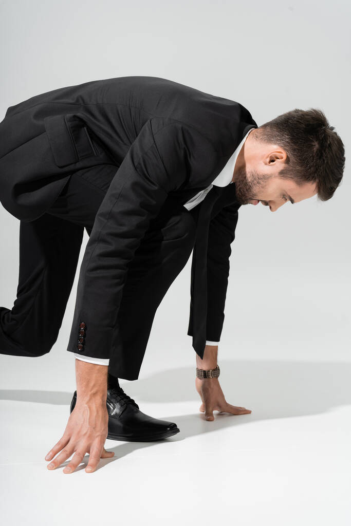 πλαϊνή άποψη του σκόπιμου επιχειρηματία σε μαύρο κοστούμι στέκεται σε χαμηλή θέση εκκίνησης σε γκρι φόντο - Φωτογραφία, εικόνα