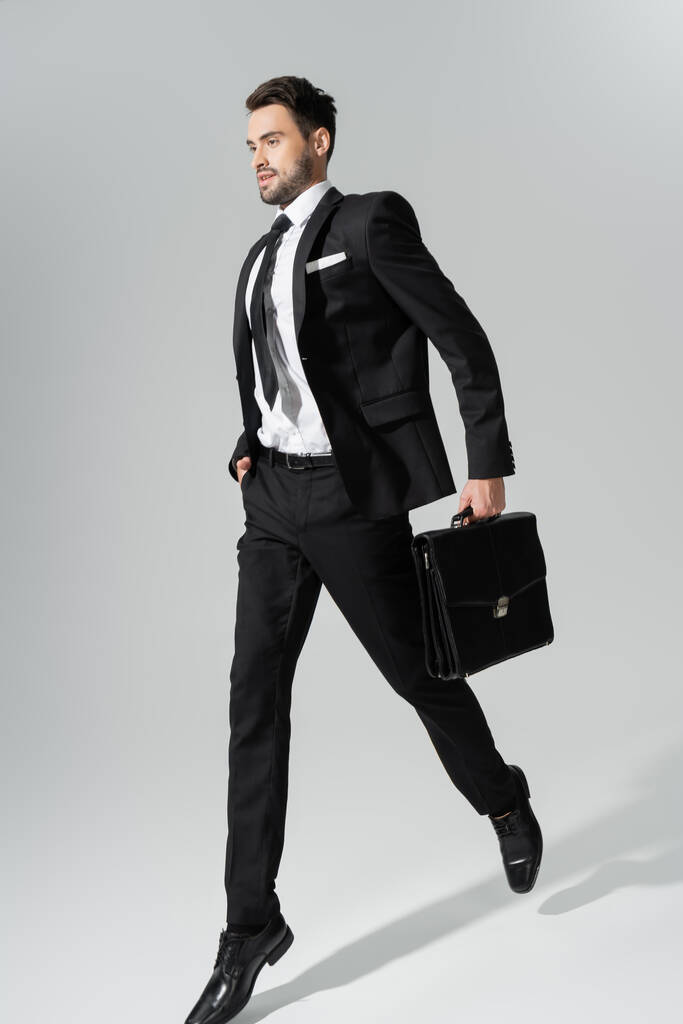 longitud completa de joven hombre de negocios en traje pantalón negro corriendo con maletín y mano en bolsillo sobre fondo gris - Foto, imagen